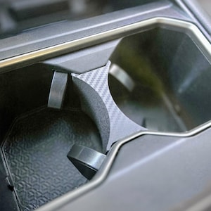 May include: Close-up of a car's cup holder with a black and gray carbon fiber pattern. The cup holder is made of plastic and has a rubberized bottom.
