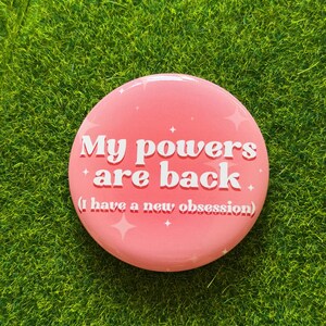 May include: A circular pink pinback button featuring white text that reads My powers are back (I have a new obsession) with small white star accents scattered across the surface. The design has a smooth finish and a vibrant pink background.