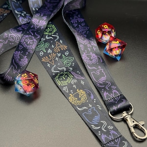 May include: Two black lanyards with colourful, glowing designs of mythical creatures and symbols. The lanyards have silver metal clasps. Two multi-coloured dice are also in the image.
