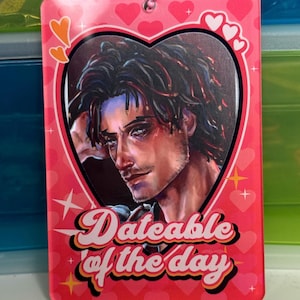 Può includere: Un portachiavi rettangolare rosa con un ritaglio a forma di cuore con un ritratto di un uomo. Il portachiavi presenta la scritta "Dateable of the day" in corsivo, con cuori e stelle come elementi decorativi.