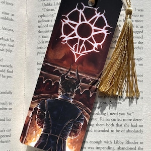 May include: A rectangular bookmark with a detailed illustration of a figure with horns, facing away from the viewer, set against a fiery background. The bookmark has a gold tassel attached to the top. A white emblem is at the top.