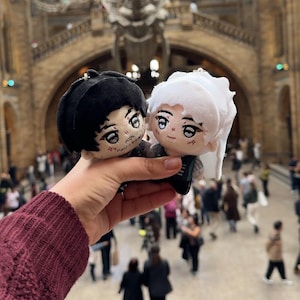 以下が含まれることがあります： 刺繍された顔の2つのぬいぐるみ人形が、博物館の背景の前に掲げられています。1つは黒髪、もう1つは白髪です。どちらの人形も顔の表情が細かく、暗い服を着ています。背景には大きな骨格と大きな階段があります。