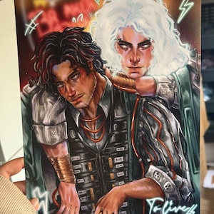 以下が含まれることがあります： 強烈な表情の2人の人物が描かれたアートプリント。一人は黒髪で真剣な表情、もう一人は白髪で最初の人物に寄り添っている。プリントは、金とティールのアクセントを持つ暗い色調で、「To live wired」というテキストが見える。