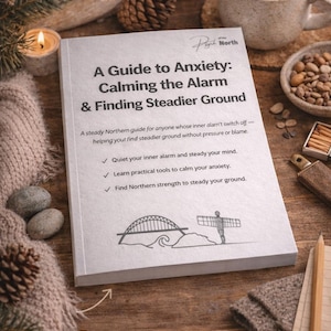 May include: A book titled "A Guide to Anxiety: Calming the Alarm & Finding Steadier Ground" is on a wooden surface. The book has a white cover with black text and illustrations. A cup of coffee, a candle, and other items are also present.