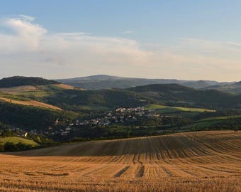 May include: A panoramic view of a rolling, grassy hillside with a small town nestled in the valley below. The hills are covered in a mix of green and brown grasses, and the sky is a clear blue.