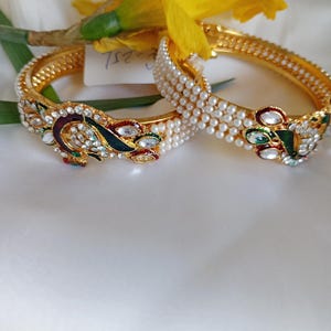 May include: Two gold bangles with white pearl accents and peacock designs. The bangles are decorated with colorful gemstones and enamel.