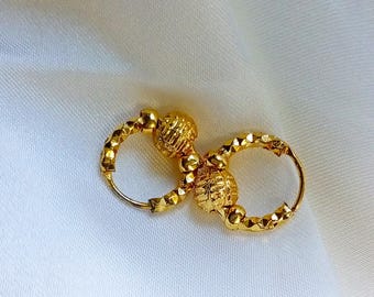 May include: A pair of gold hoop earrings with textured ball accents. The earrings are on a white fabric background.