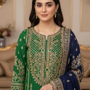 May include: A green embroidered dress with gold detailing and a navy blue shawl. The dress features intricate gold patterns on the bodice and sleeves. A gold necklace with square pendants and matching earrings complete the look. The outfit is suitable for special occasions.