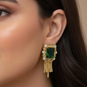 May include: Gold-toned earrings with a rectangular emerald-green stone center, surrounded by smaller clear stones. The earrings have a chain fringe design hanging below the main stone. The earrings are set against a blurred background.
