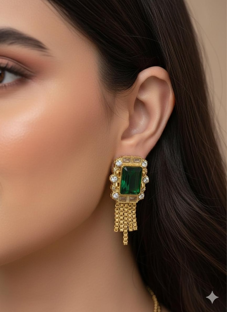 May include: Gold-toned earrings with a rectangular emerald-green stone center, surrounded by smaller clear stones. The earrings have a chain fringe design hanging below the main stone. The earrings are set against a blurred background.