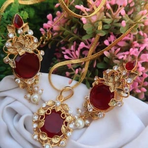 May include: A gold-coloured jewellery set comprising a necklace and earrings. Each piece features a large, round, deep red stone, surrounded by gold filigree and small, clear crystals. The set includes faux pearls.