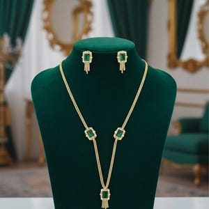 May include: A gold-toned jewelry set featuring a necklace and matching earrings. The necklace has a series of emerald-colored, square-cut stones. The earrings mirror the design with similar emerald-colored stones. The jewelry is displayed on a green velvet bust.