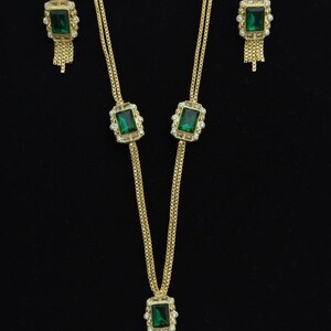 May include: Gold-toned jewelry set featuring a necklace and matching earrings. The necklace has a rectangular green gemstone pendant with a fringe, and the earrings have a similar design. The set is displayed against a black background.