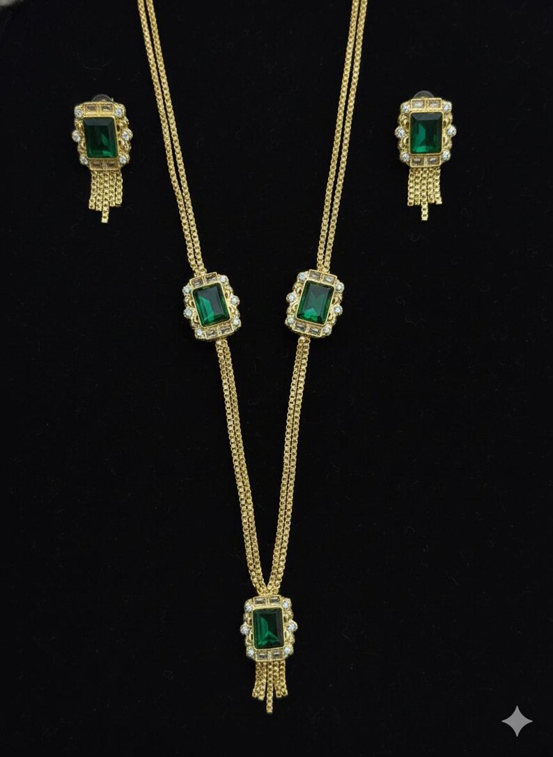 May include: Gold-toned jewelry set featuring a necklace and matching earrings. The necklace has a rectangular green gemstone pendant with a fringe, and the earrings have a similar design. The set is displayed against a black background.