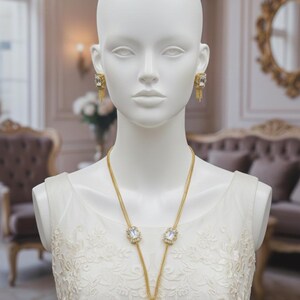 May include: A gold-toned necklace and matching earrings set on a mannequin. The jewelry features large, square, clear gemstones and delicate chain details. The mannequin is wearing a white, lacy dress, and the background suggests a luxurious setting.