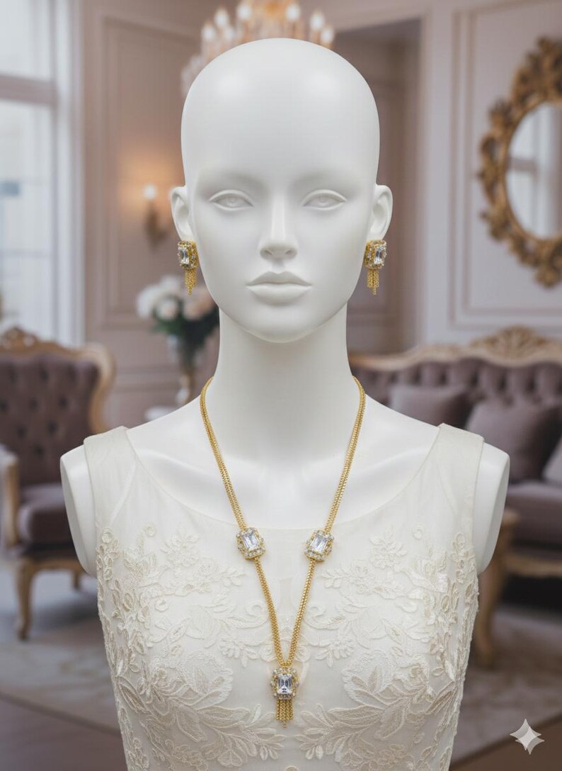 May include: A gold-toned necklace and matching earrings set on a mannequin. The jewelry features large, square, clear gemstones and delicate chain details. The mannequin is wearing a white, lacy dress, and the background suggests a luxurious setting.