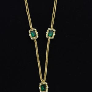 May include: Gold-toned necklace with emerald green rectangular stones and clear accent stones. The necklace has a Y-shaped design with a tassel at the bottom. The background is black.