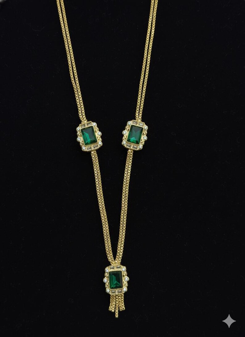 May include: Gold-toned necklace with emerald green rectangular stones and clear accent stones. The necklace has a Y-shaped design with a tassel at the bottom. The background is black.