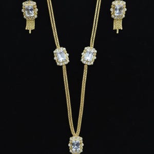 May include: A gold-toned jewelry set featuring a necklace and matching earrings. The necklace has a rectangular pendant with a large, clear stone and a fringe of small chains. The earrings mirror the pendant design.