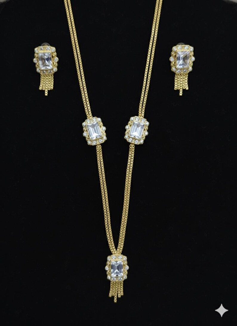 May include: A gold-toned jewelry set featuring a necklace and matching earrings. The necklace has a rectangular pendant with a large, clear stone and a fringe of small chains. The earrings mirror the pendant design.