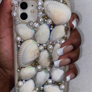 May include: A beige phone case decorated with white seashells, faux pearls, and gold beads. The case has a cluster of shells around the camera and is decorated with iridescent glitter. The case is designed to protect the phone.