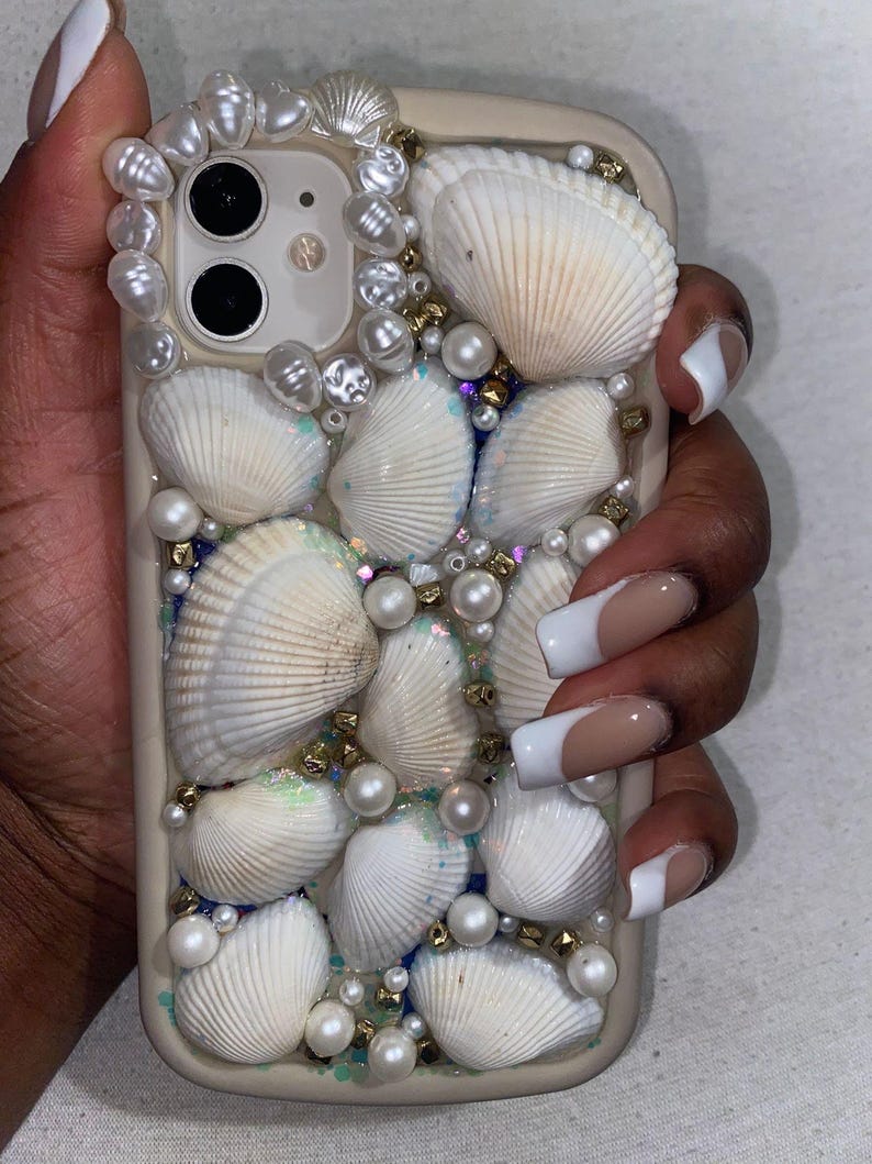 May include: A beige phone case decorated with white seashells, faux pearls, and gold beads. The case has a cluster of shells around the camera and is decorated with iridescent glitter. The case is designed to protect the phone.