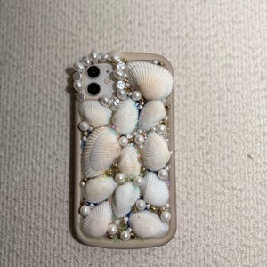 Può includere: Una custodia per telefono beige ornata di conchiglie bianche, perle e perline dorate. La custodia presenta un gruppo di conchiglie e perle attorno alla fotocamera. Il design crea un'estetica balneare e costiera.