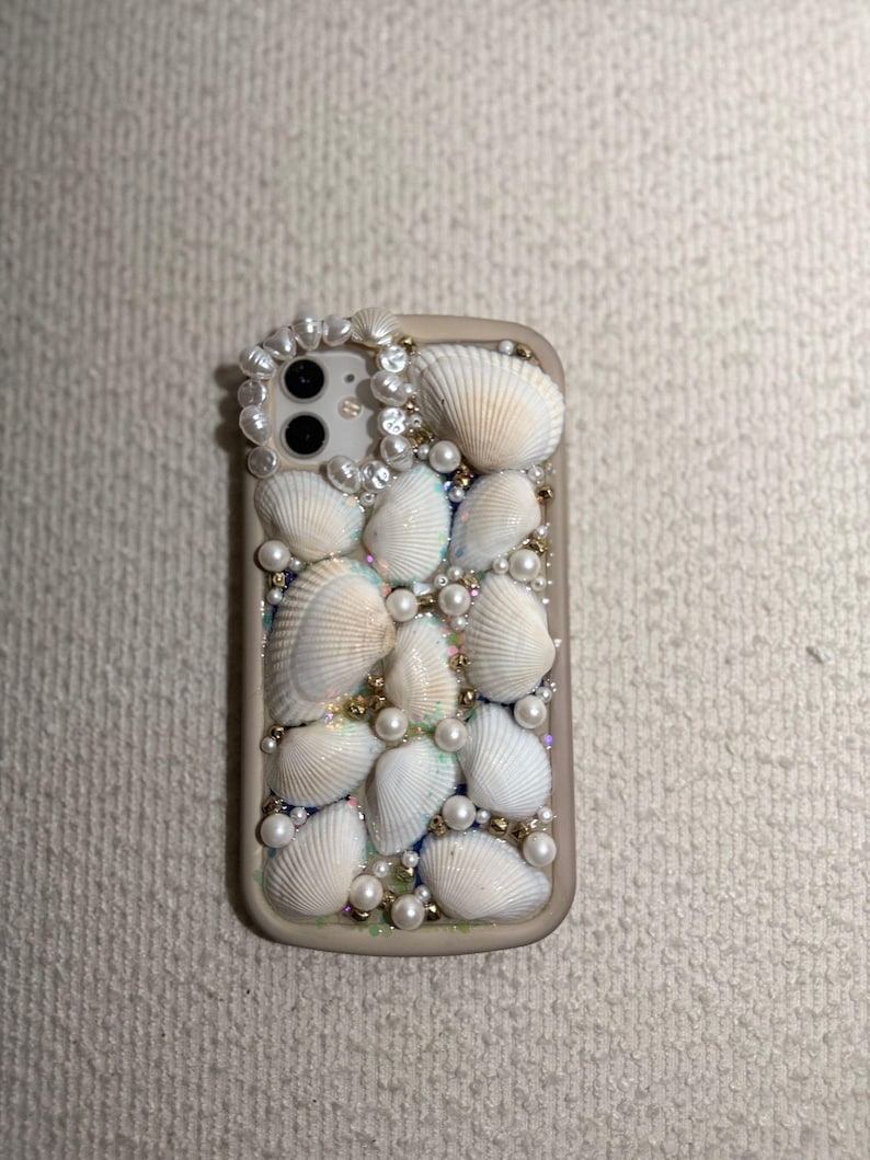 May include: A beige phone case decorated with white seashells, pearls, and gold beads. The case has a cluster of seashells and pearls around the camera. The design creates a beachy, coastal aesthetic.