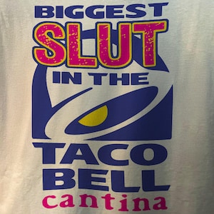 Puede incluir: Una camiseta gris claro con las palabras "BIGGEST SLUT IN THE TACO BELL cantina" en letras azules, rosas y amarillas. El logotipo de Taco Bell también aparece en azul y amarillo.