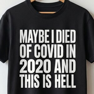 Op de afbeelding: Zwart T-shirt met de witte tekst "MAYBE I DIED OF COVID IN 2020 AND THIS IS HELL." Het shirt hangt aan een houten hanger. De achtergrond toont een witte muur, planten en een bank.