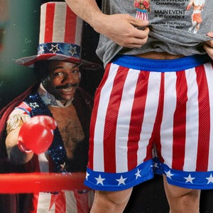 May include: Red and white striped mesh shorts with a blue waistband and star accents. The shorts are part of a boxing-themed outfit, with a matching Uncle Sam hat and a boxing ring in the background.