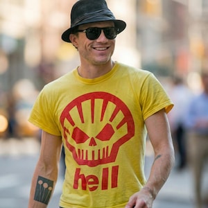 Könnte beinhalten: Ein gelbes T-Shirt mit einem roten Totenkopf-Motiv und dem Wort "hell" in Rot. Die Person trägt einen schwarzen Fedora-Hut und eine Sonnenbrille. Ein Tattoo mit drei Kreisen ist auf dem Arm zu sehen.