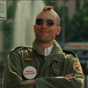 Puede incluir: Un hombre con gafas de sol de aviador y una chaqueta verde de estilo militar con parches y un botón que dice "We Are The People". El hombre tiene un peinado mohawk y está sonriendo.