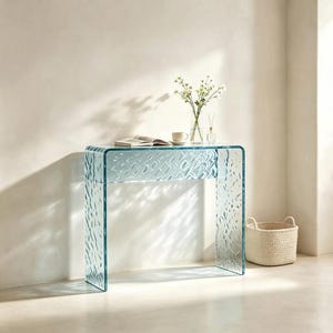 Könnte beinhalten: Ein hellblauer Glaskonsolentisch mit einer texturierten, gewellten Oberfläche. Der Tisch hat ein Wasserfall-Design mit geschwungenen Kanten. Darauf stehen eine klare Glasvase mit weißen Blumen, eine weiße Keramiktasse und ein aufgeschlagenes Buch.