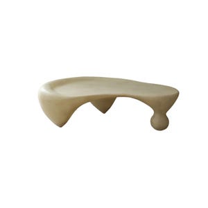 May include: A beige, organically shaped coffee table with a smooth, curved surface. The table has three unique leg supports, including a rounded sphere and two triangular shapes. The table is approximately 152 cm long.