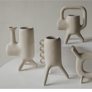 Może przedstawiać: Kolekcja kremowych wazonów ceramicznych o unikalnych, abstrakcyjnych kształtach. Jeden wazon ma uchwyt, inny szereg okrągłych akcentów, a inne mają wydłużone formy. Wazony są prezentowane na białej powierzchni.