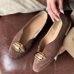 以下が含まれることがあります： ゴールドのバックルが付いた、ブラウンのスエードフラットシューズ。靴はポインテッドトゥで、ローヒールです。