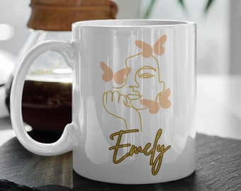 May include: White ceramic coffee mug with a gold line drawing of a woman's face with butterflies around her. The name "Emely" is written in gold script below the drawing.