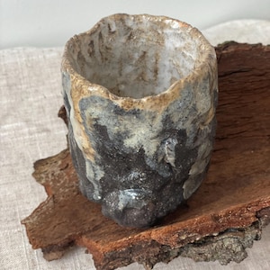 Op de afbeelding: Een cilindervormige keramische beker met een ruwe, onregelmatige rand en een textuurrijk oppervlak. De buitenkant heeft een donkergrijze en zwarte basis met druipende crème- en beige glazuuraccenten. De binnenkant is licht, gespikkeld gebroken wit.