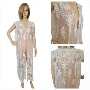 May include: An ivory lace duster coat, sleeveless and floor-length. The open front reveals intricate floral and leaf embroidery. The sheer fabric allows the undergarments to be seen. The duster is displayed on a mannequin.