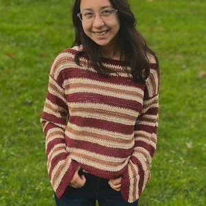 Puede incluir: Un suéter de ganchillo a rayas horizontales en burdeos, beige y crema. El suéter tiene mangas largas y un corte holgado. La persona lleva vaqueros azul oscuro. El fondo es hierba verde.