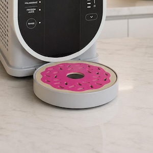 May include: A light gray kitchen appliance with a black control panel featuring text labels for Milkshake, Frozen Juice, and Rinse. A circular, light gray drip tray with a pink donut-shaped insert featuring small dash-like cutouts sits in front.