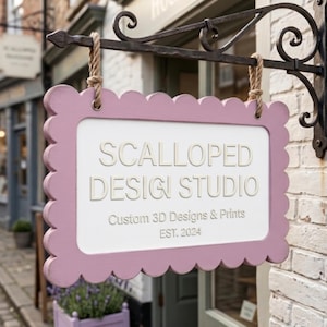 Puede incluir: Un letrero rectangular colgante con un borde festoneado de color rosa polvoriento y un panel central blanco. El texto dice SCALLOPED DESIGN STUDIO, Custom 3D Designs & Prints y EST. 2024. El letrero cuelga de dos cuerdas gruesas de un soporte de metal negro.