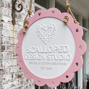 Può includere: Un'insegna rotonda con bordo smerlato, cornice rosa e centro bianco, sospesa tramite due corde spesse a un supporto in metallo nero. L'insegna presenta un'icona a forma di cuore bianco e il testo SCALLOPED DESIGN STUDIO, DIGITAL & CUSTOM CREATIONS, EST. 2026.