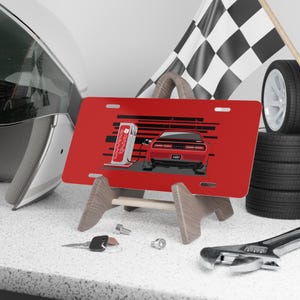 Puede incluir: Placa de matrícula roja con una ilustración estilizada de un coche rojo en una estación de carga. La placa se muestra en un soporte de madera, con una bandera a cuadros y neumáticos en el fondo. También se ven un casco, herramientas y llaves.