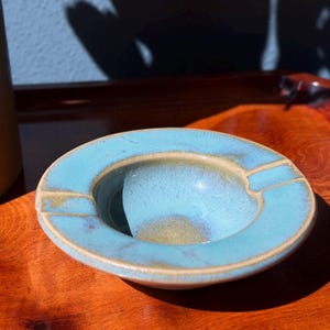 May include: A round, ceramic ashtray with a turquoise glaze. The ashtray has a circular design with a raised rim and two rectangular rests. The interior is a lighter shade of turquoise with a glossy finish. The ashtray sits on a wooden surface.