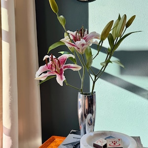 Puede incluir: Un jarr&oacute;n plateado contiene lirios rosas y blancos con hojas y capullos verdes. El jarr&oacute;n est&aacute; sobre una pila de libros, junto a un cenicero de m&aacute;rmol blanco con dos cajas de cerillas. Un espejo decorativo cuelga de la pared turquesa.