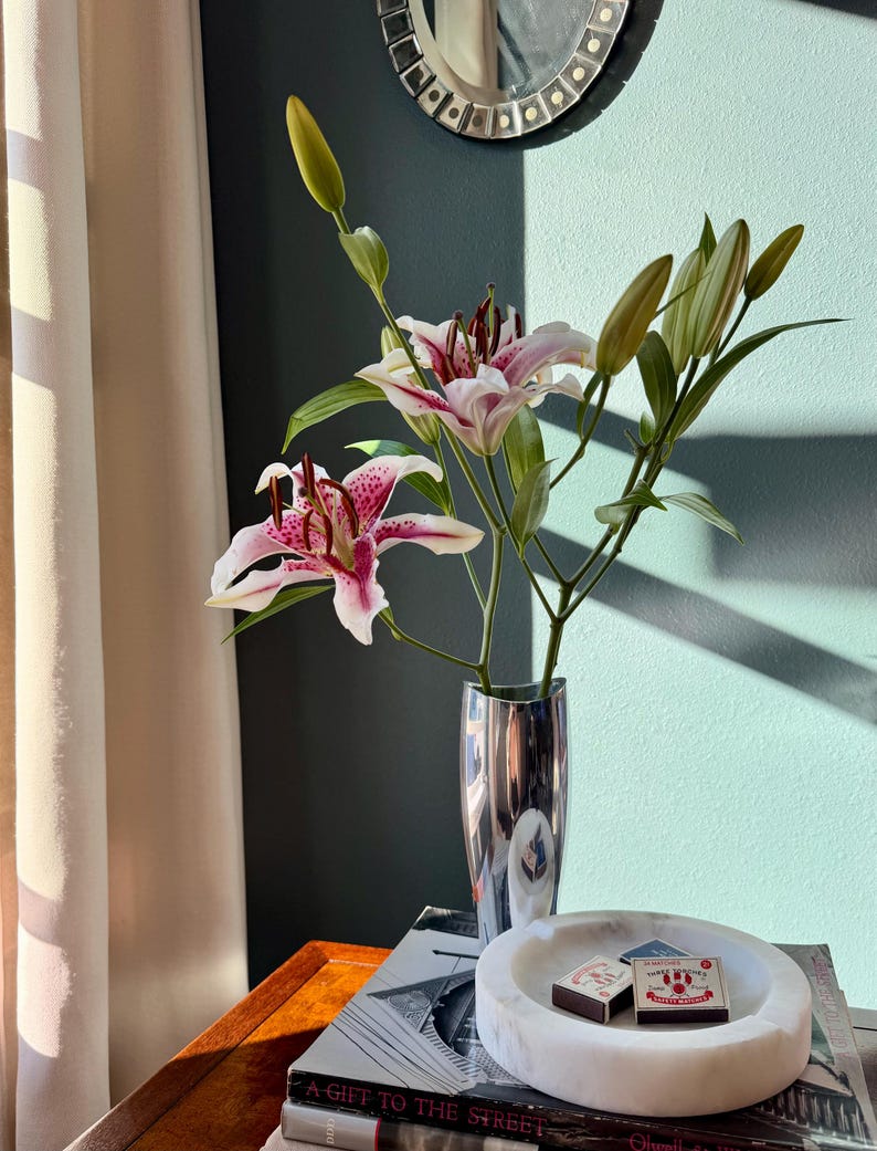 Puede incluir: Un jarr&oacute;n plateado contiene lirios rosas y blancos con hojas y capullos verdes. El jarr&oacute;n est&aacute; sobre una pila de libros, junto a un cenicero de m&aacute;rmol blanco con dos cajas de cerillas. Un espejo decorativo cuelga de la pared turquesa.