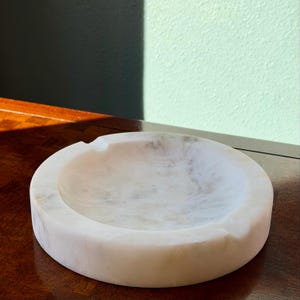 Puede incluir: Un cenicero redondo de m&aacute;rmol blanco con un cuenco poco profundo y un borde curvo. El cenicero tiene un patr&oacute;n veteado natural y una peque&ntilde;a muesca para los cigarrillos. Un art&iacute;culo decorativo para el hogar.