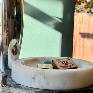 Puede incluir: Un cenicero de m&aacute;rmol blanco con un dise&ntilde;o circular, que contiene dos tarjetas peque&ntilde;as y una caja de cerillas. La caja de cerillas dice "THREE TORCHES" y "SAFETY MATCHES". Tambi&eacute;n se ven un jarr&oacute;n plateado y un libro.